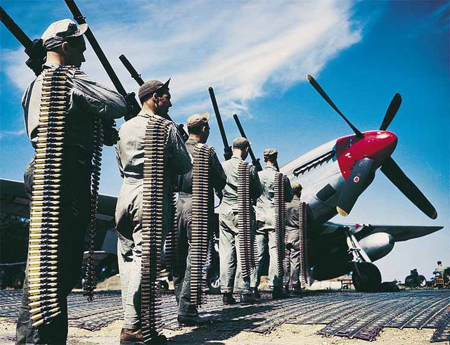 Armorers carry six Browning AN/M2 .50-caliber machine guns and belts of linked ammunition to a parked P-51 Mustang. (U.S. Air Force)