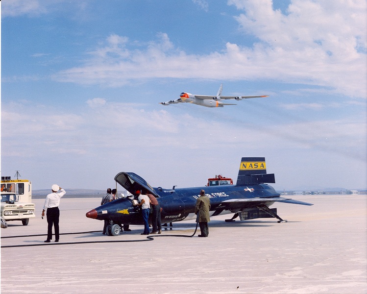 A North American Aviation support crew deactivates X-15 56-6671 on Rogers Dry Lake after a flight, while the mothership, NB-52A Stratofortress 52-003 flies overhead. (NASA)