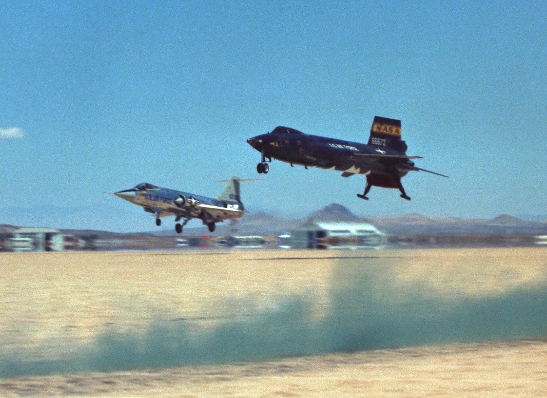 The number three North American Aviation X-15 rocketplane, 56-6672, just before touchdown on Rogers Dry Lake. A Lockheed F-104 Starfighter chase plane escorts it. The green smoke helps the pilots judge wind direction and speed. Frost on the X-15's belly shows residual propellants in the tanks. (NASA)