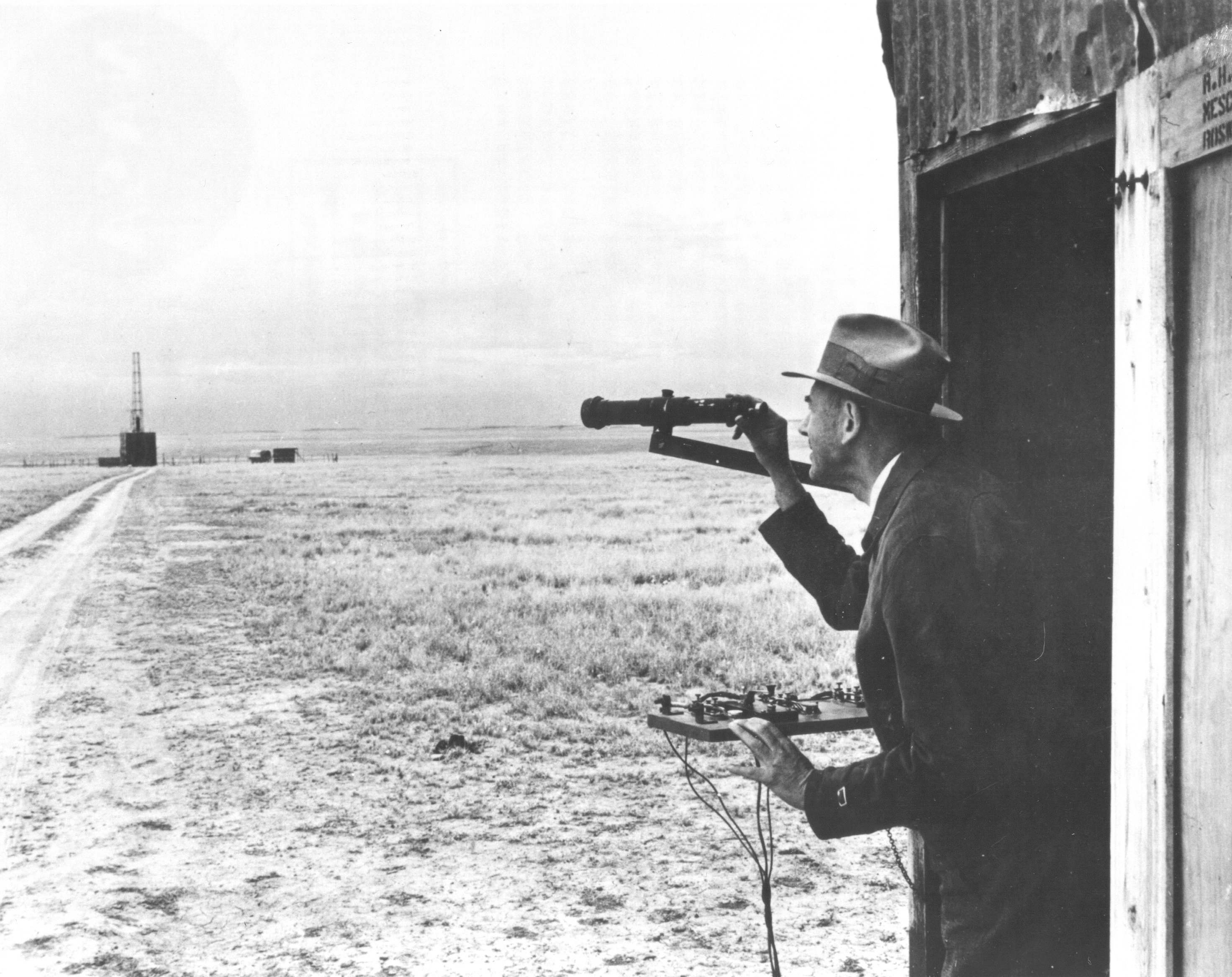 "Dr. Robert H. Goddard observes the launch site from his launch control shack while standing by the firing control panel. From here he can fire, release, or stop testing if firing was unsatisfactory. Firing, releasing, and stop keys are shown on panel. The rocket is situated in the launch tower." (NASA) 
