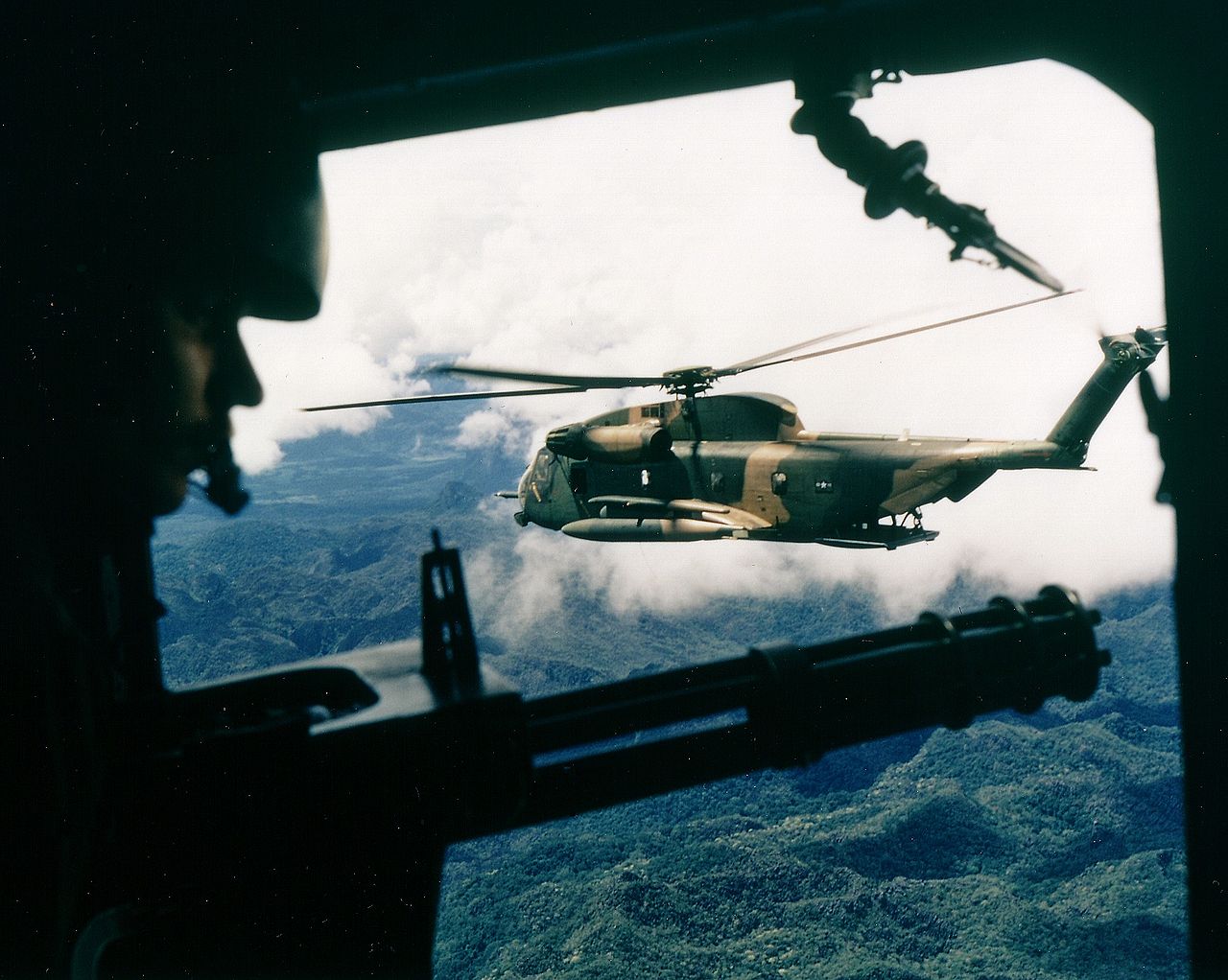 A gunner looks over a General Electric GAU2/A minigun, while his aircraft flies formation with a Sikorsky HH-53C Super Jolly Green Giant over Southeast Asia. (U.S. Air Force)